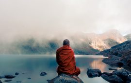 A monk from back sitting on the stone near the water in the background of foggy mountains.